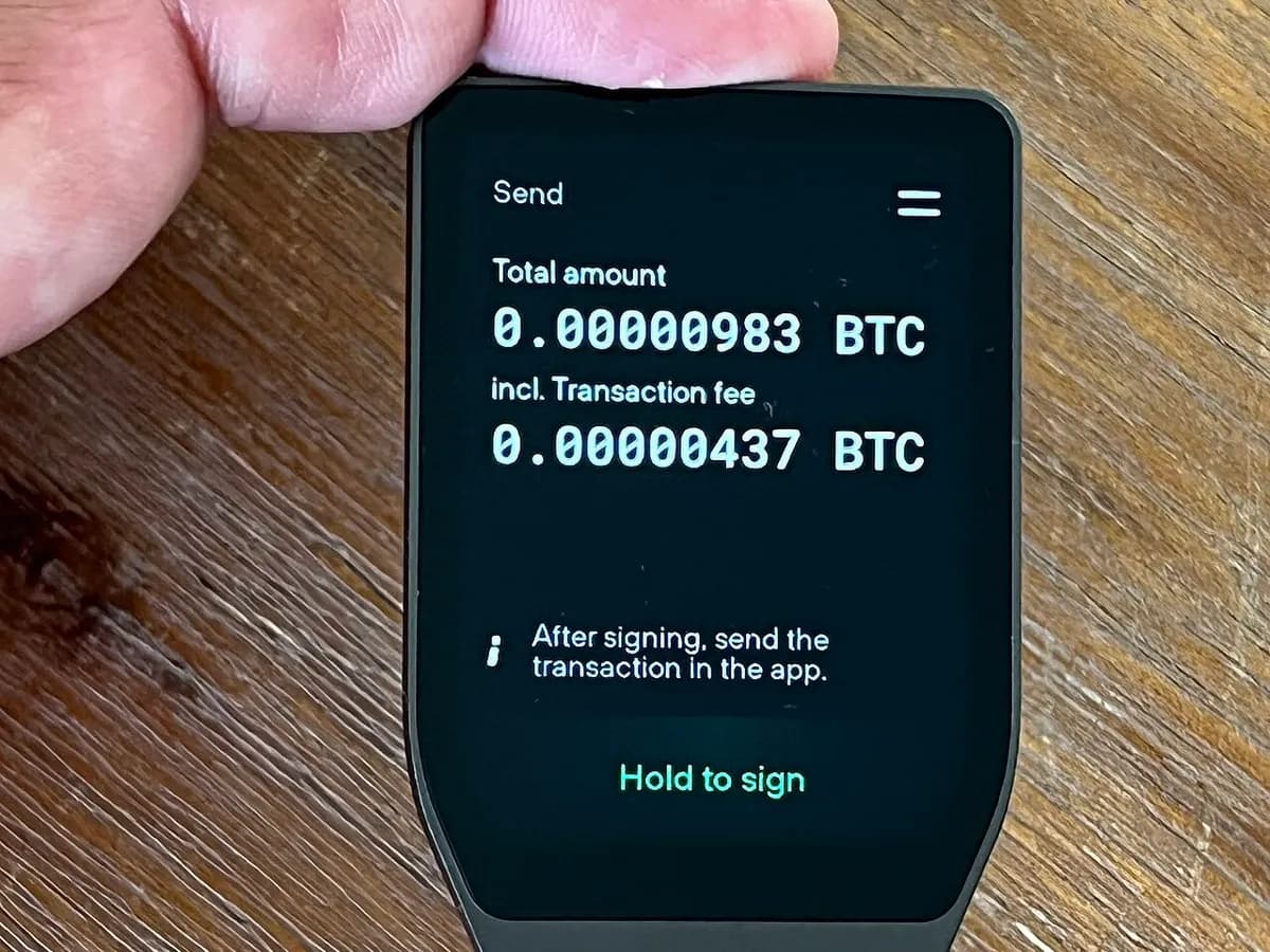 A hardware wallet held in one hand, showing a transfer to confirm on its own screen with the words Hold to sign.