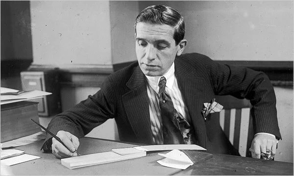 Black-and-white photograph of Charles Ponzi seated at a desk, writing, in a suit and tie.